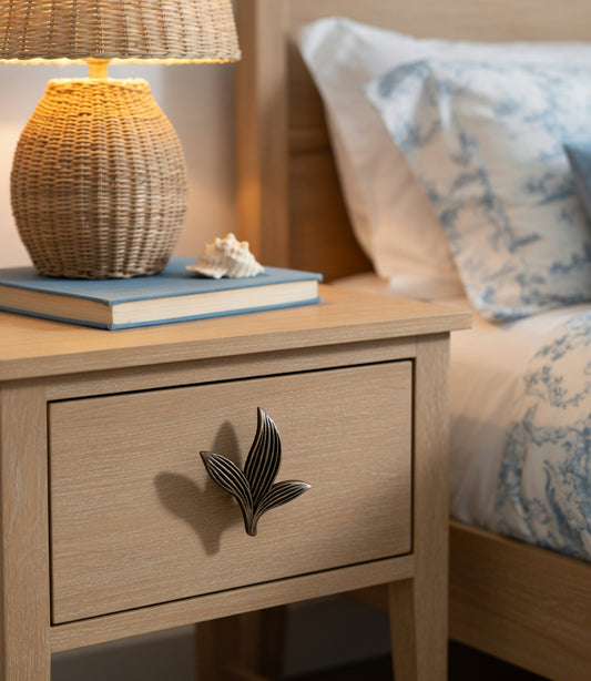 Wooden nightstand with bronze Sera knob, wicker lamp and decorative items next to a bed with patterned bedding.