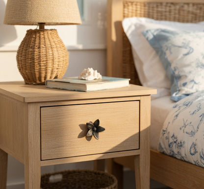 Wooden nightstand with petite flower knob, a lamp and books next to a bed with floral bedding.