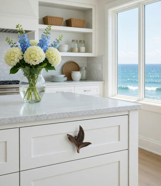 Kitchen with white cabinets, bronze Sera leaf knob, marble countertop, and a vase of flowers by a window with ocean view.