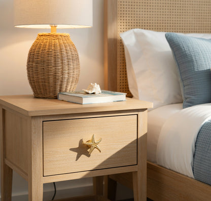 Wooden nightstand with wicker lamp and books next to a bed with white and blue bedding.